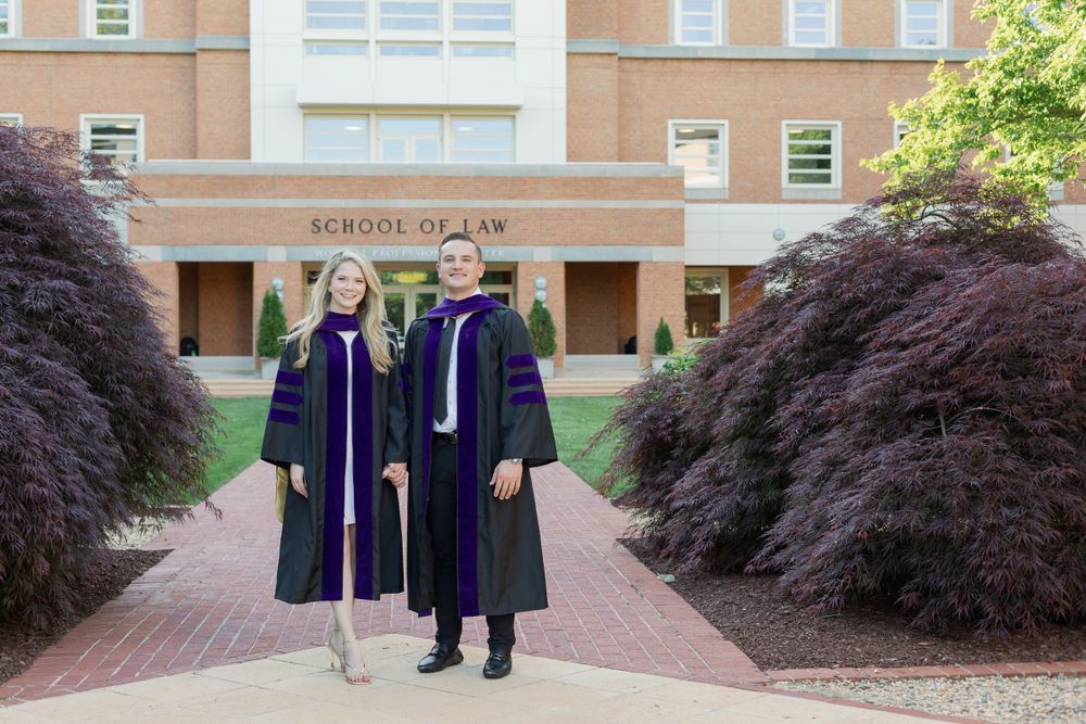 Graduates in purple and black academic regalia stand on brick pathway at law school entrance.