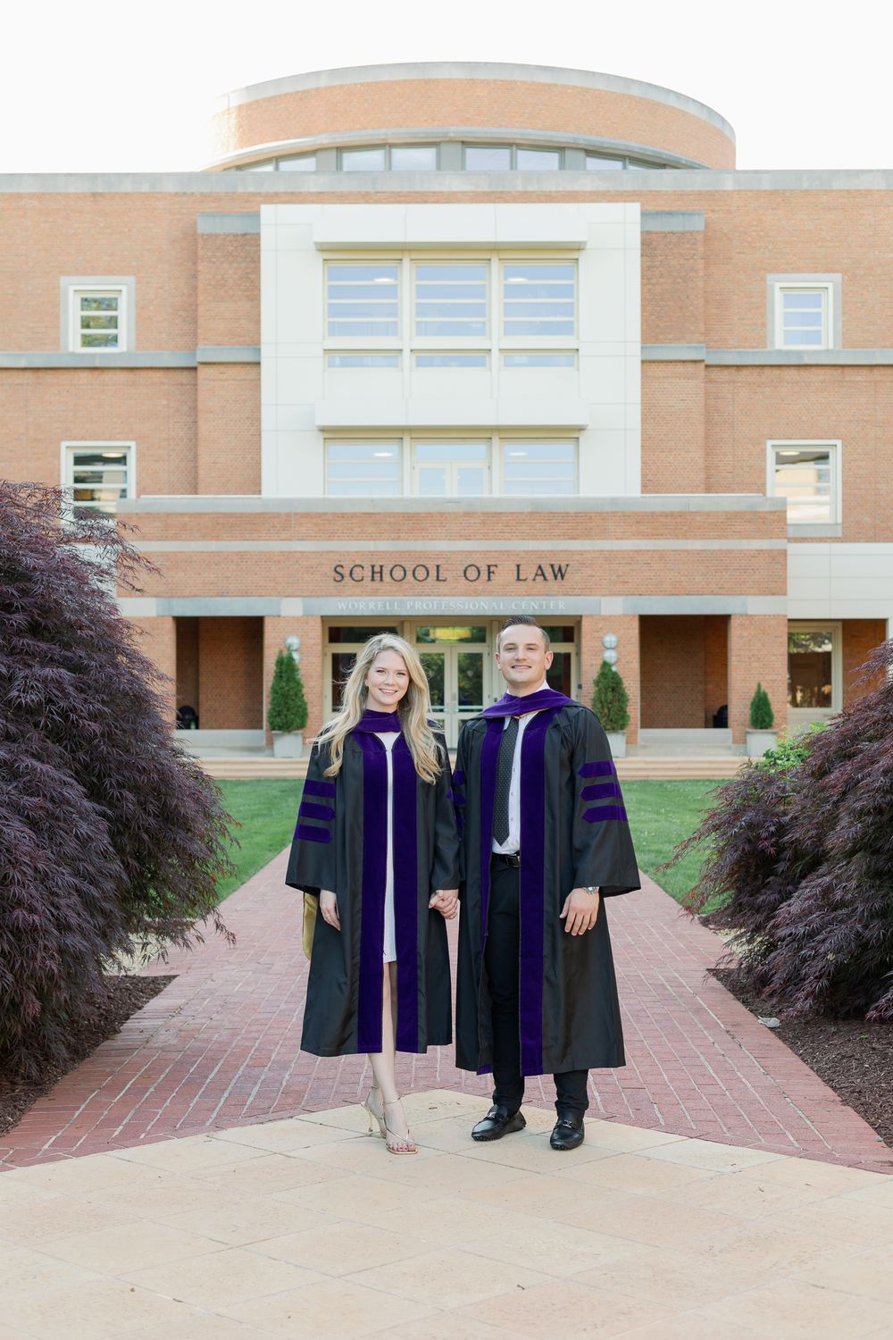Two graduates in academic regalia pose in front of School of Law building entrance.
