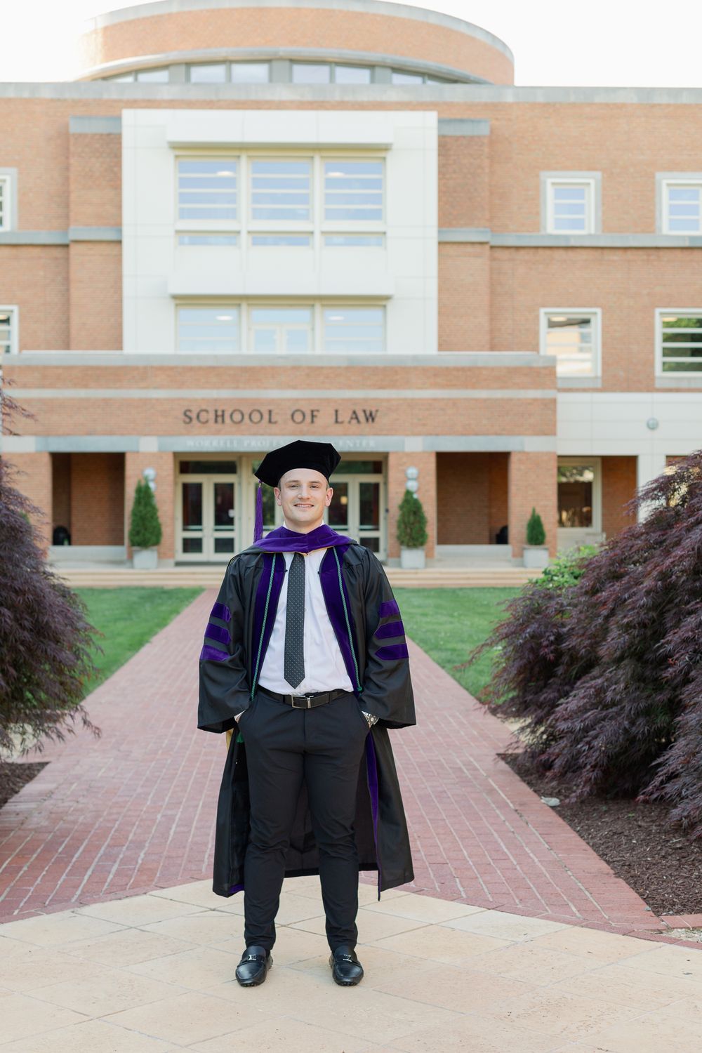 Graduate in academic regalia poses on brick pathway in front of School of Law building.
