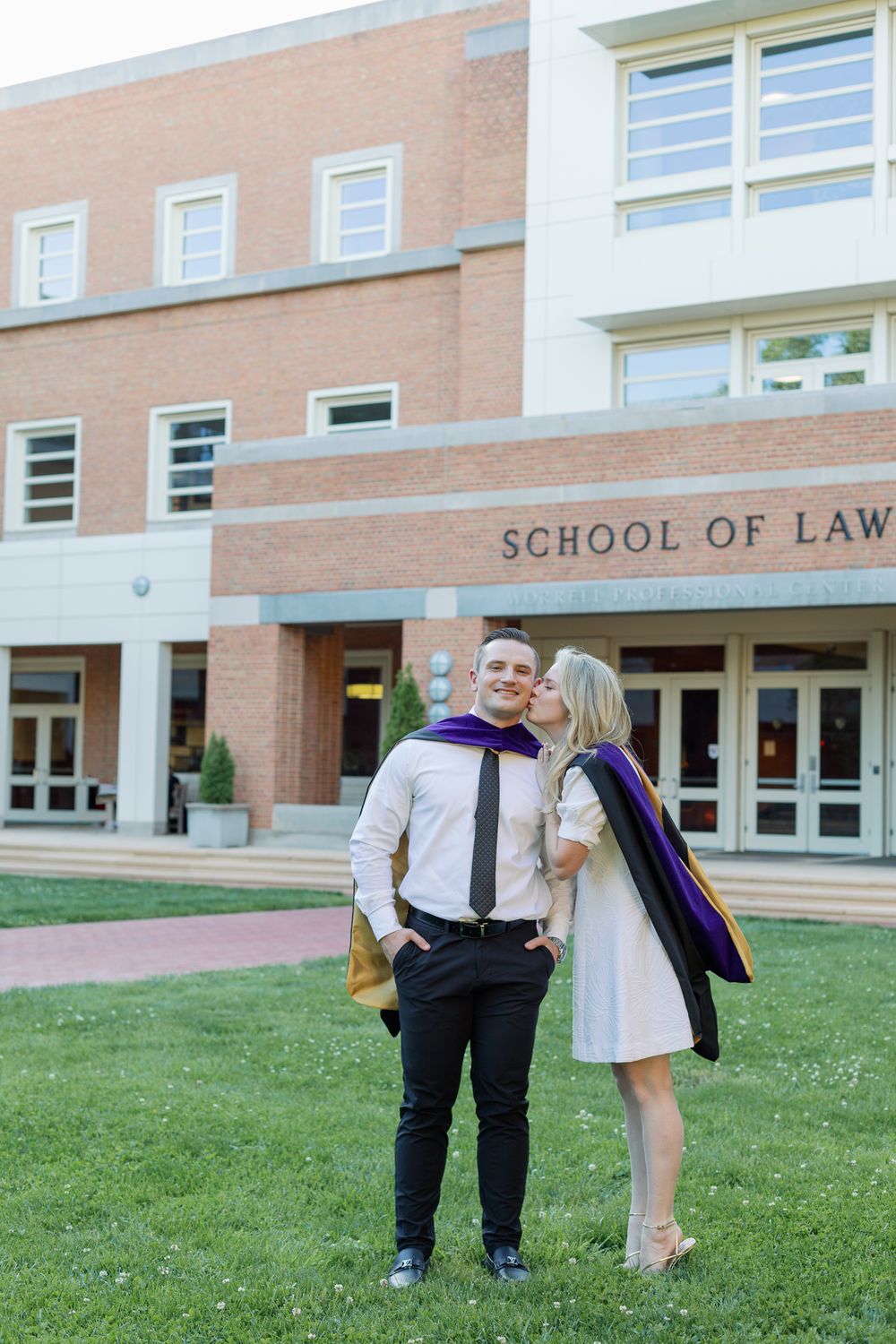 Two graduates in formal attire pose together on the lawn outside a School of Law building.