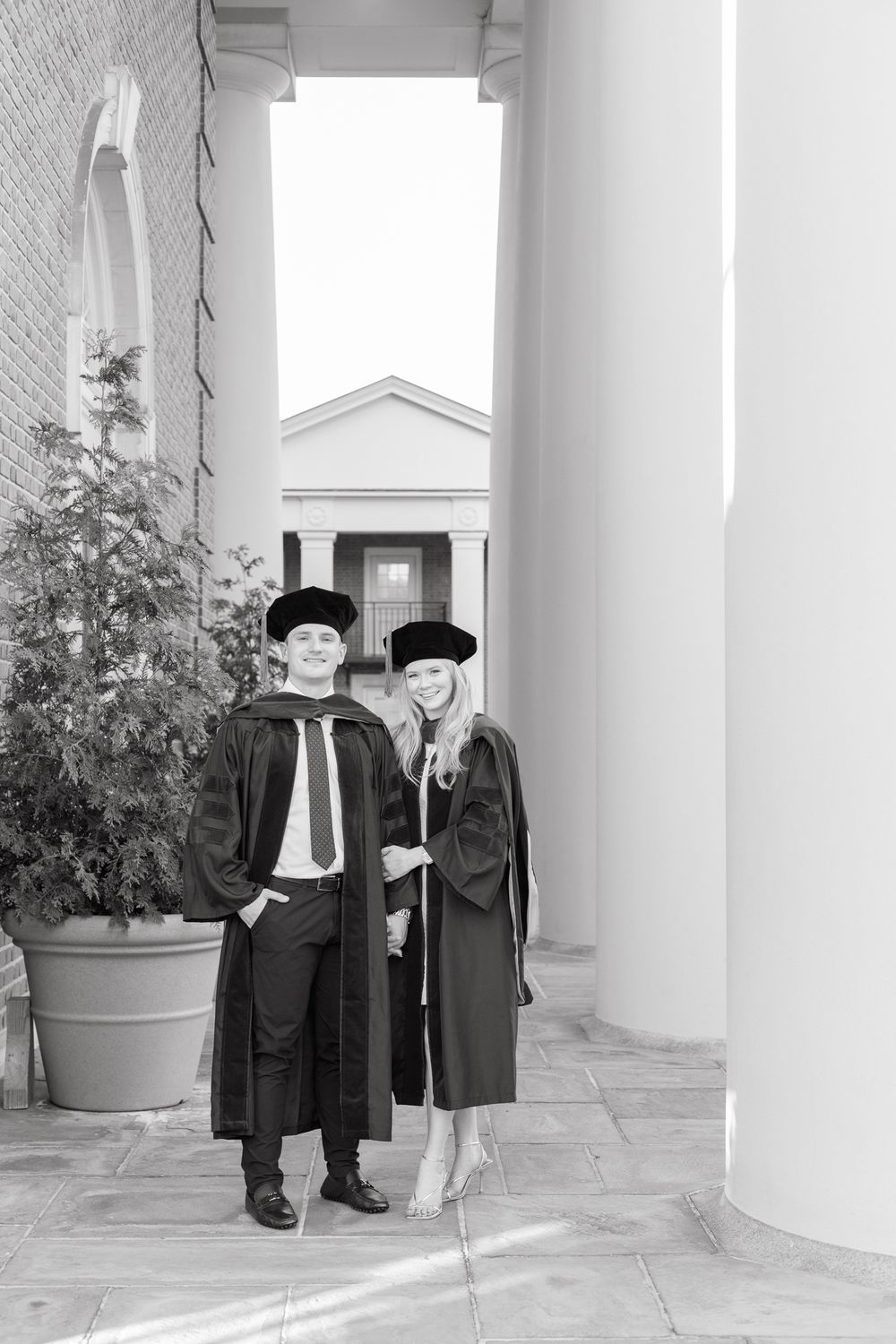 Two graduates in black caps and gowns pose between white columns in black and white.