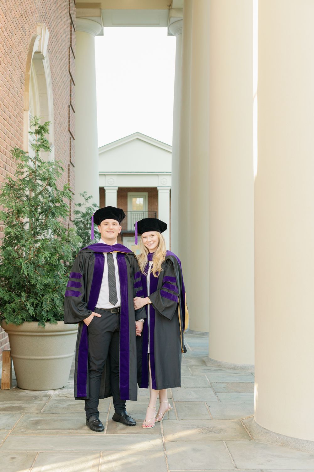 Graduates in purple and black academic regalia stand between architectural columns.