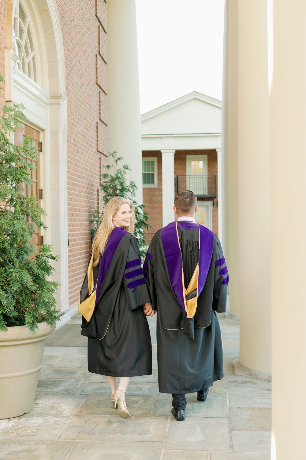Graduates in black and purple academic robes walk through a columned walkway.