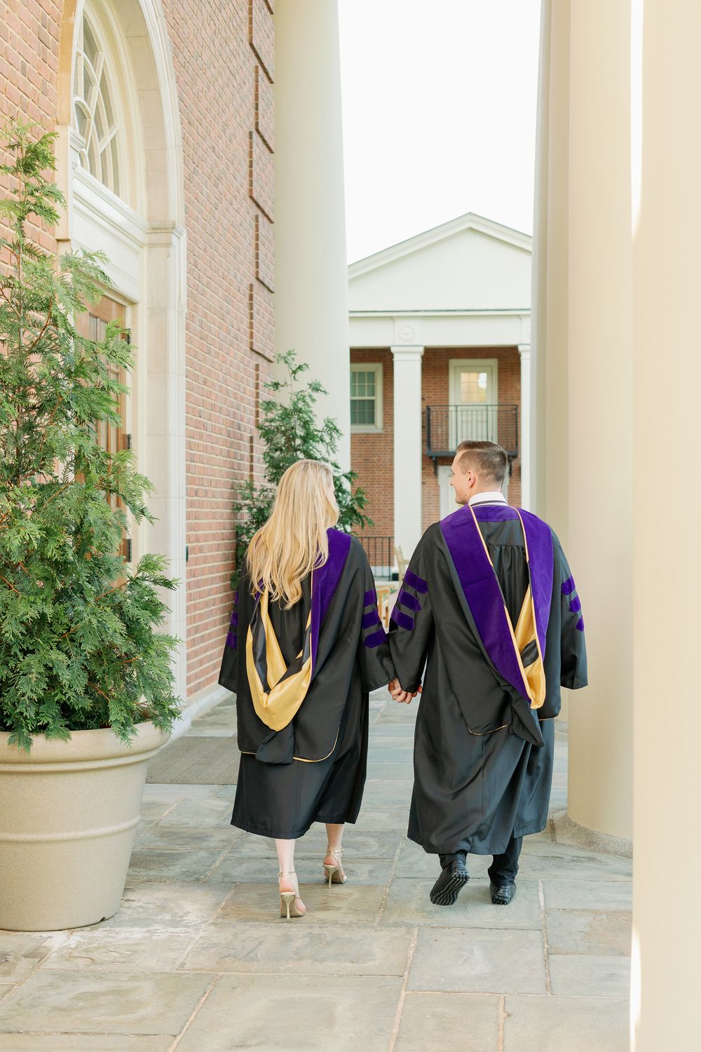 Law school graduates holding hands walk through a columned courtyard in their regalia.