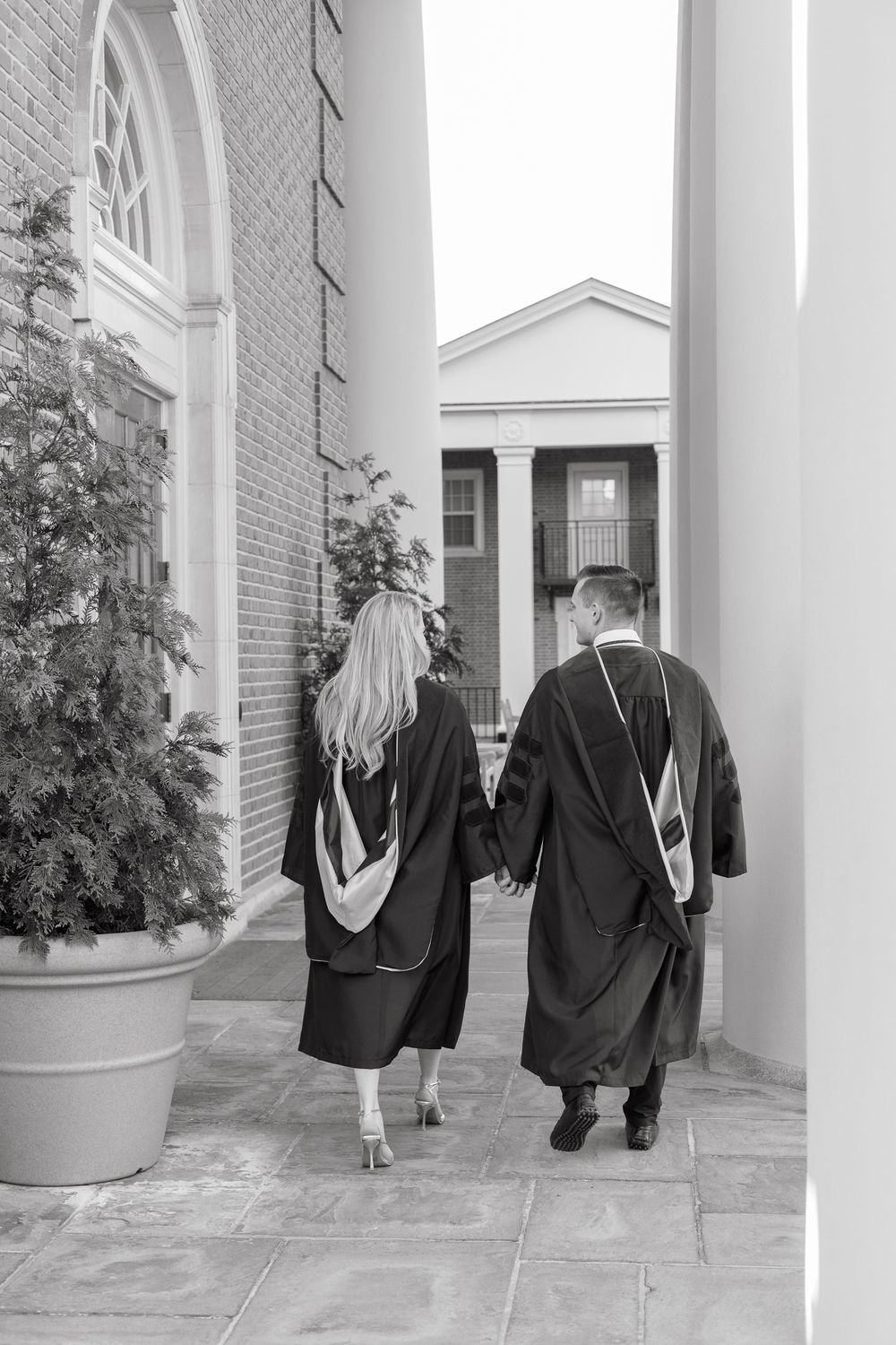 Graduates in black robes walk hand in hand through a colonnade in black and white.