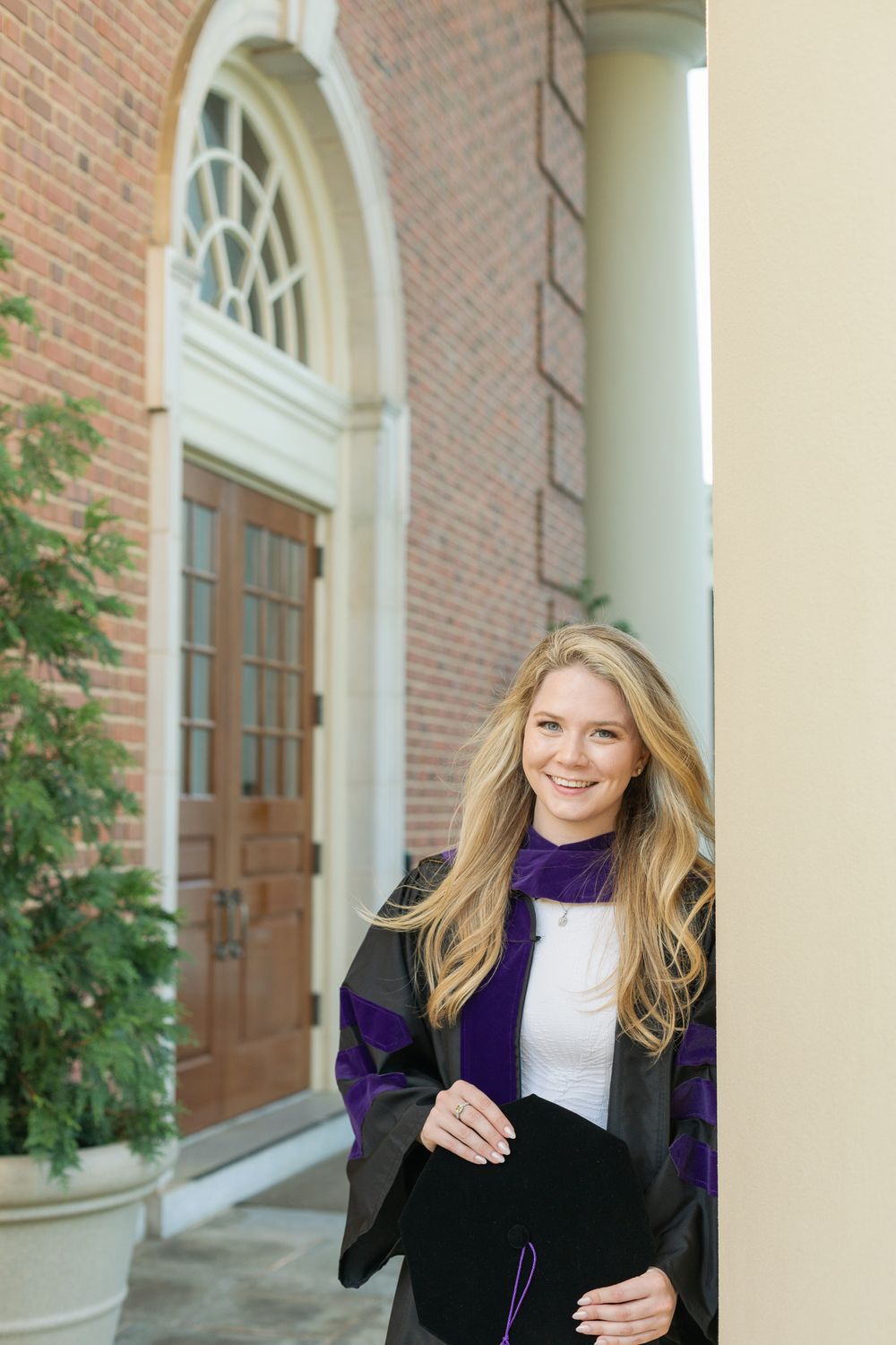 Graduate wearing black and purple academic regalia poses in front of brick building with white columns.