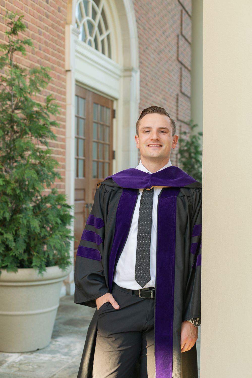 Graduate in black cap and gown with purple stole smiles in front of collegiate architecture.