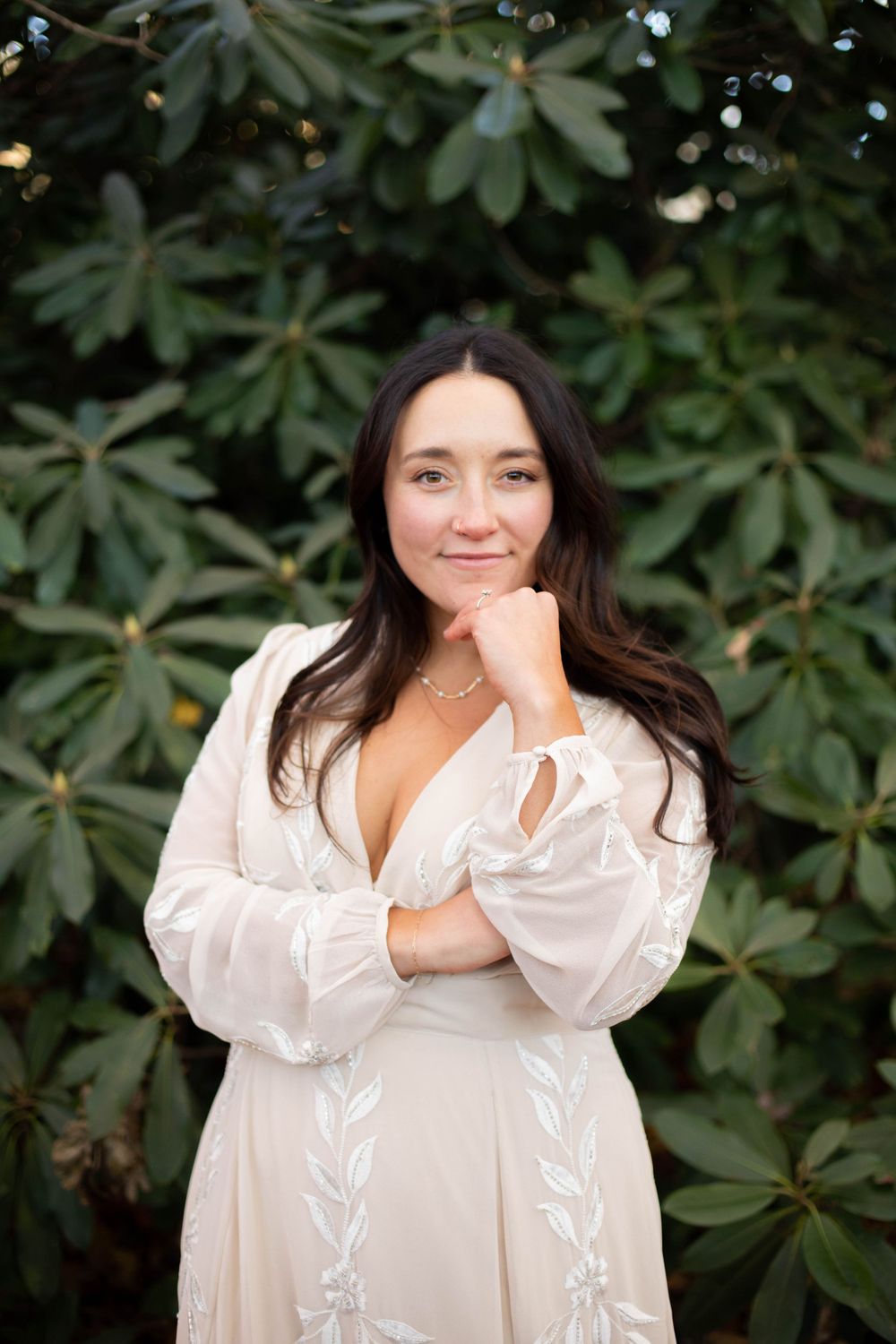Person in a white embroidered dress poses against green foliage background.