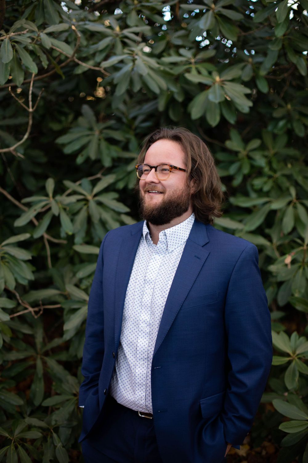 Person in navy suit and polka dot shirt poses against green foliage backdrop.