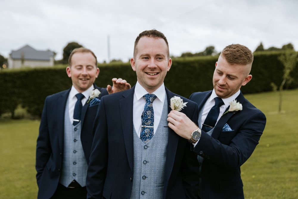 Group of groomsmen in matching suits pose outdoors on a green lawn.