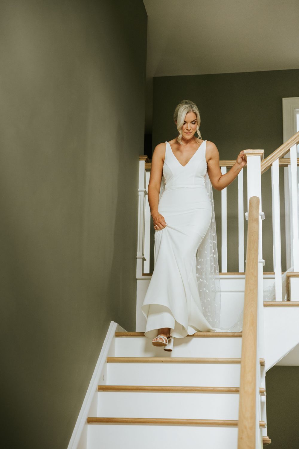 Bride in white dress descends elegant staircase with wooden railing.