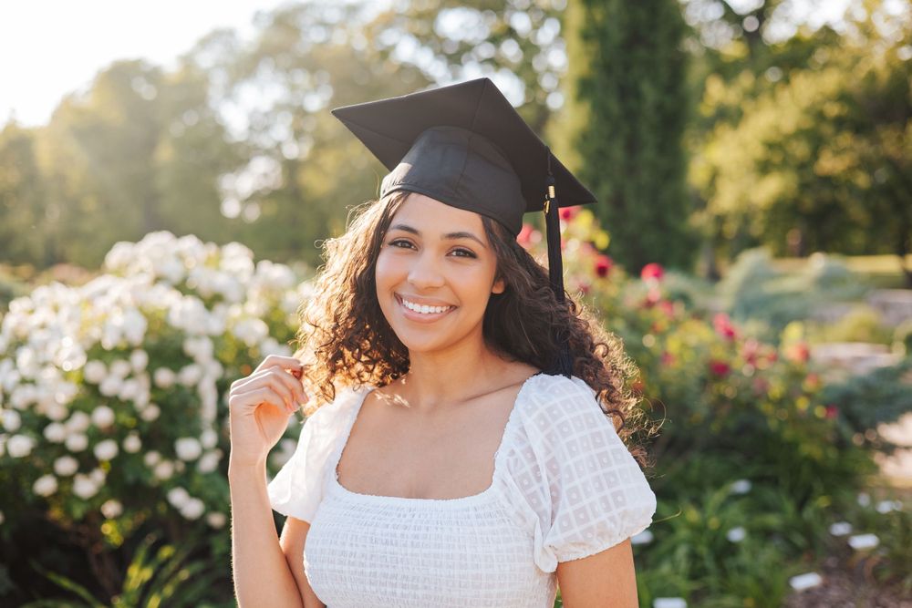 Graduate in cap and white dress smiling in garden with flowers.