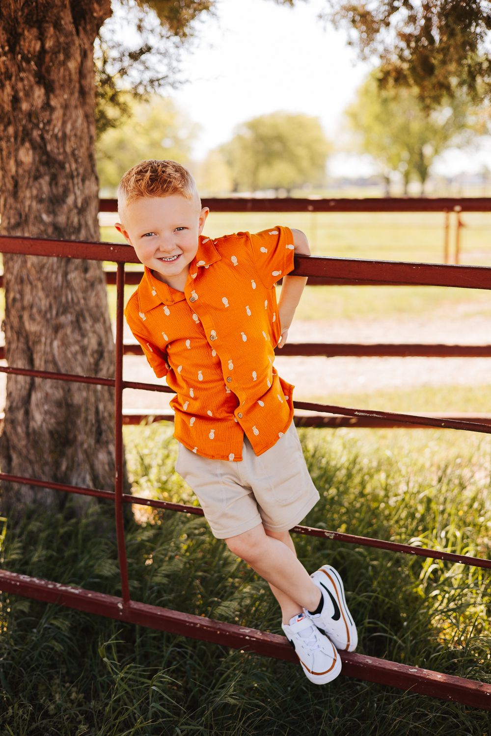 Child in bright orange shirt playing on red metal fence rails in sunny outdoor setting.