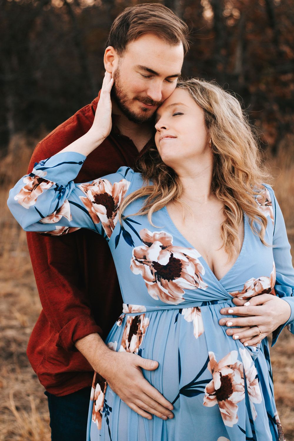 Romantic couple embracing in field wearing blue floral dress and burgundy top at sunset.