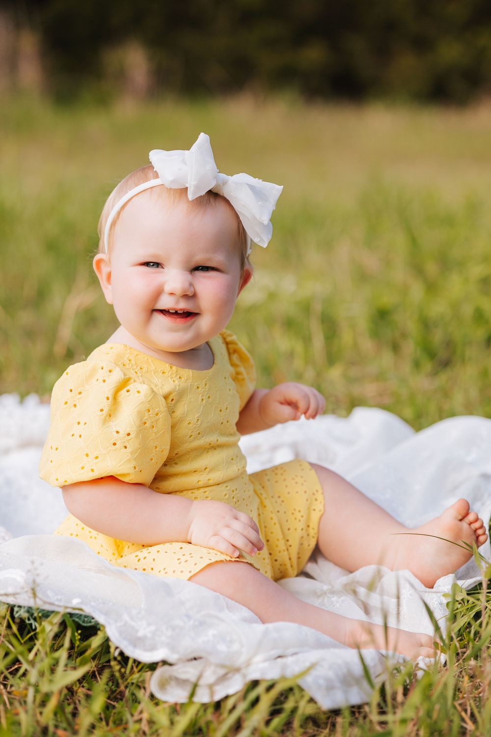 Baby in yellow eyelet dress and white bow lying on blanket in grass during outdoor photo session.