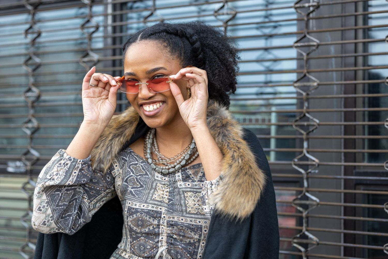 Stylish Black woman smiling while adjusting pink tinted sunglasses outdoors wearing patterned dress and fur wrap.