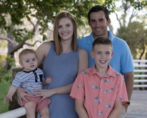 A happy family of four poses together outdoors on a sunny day with white fencing and trees in the background.