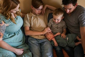 A group of people sit closely together on a bed while looking at newborn in younger brother's lap.