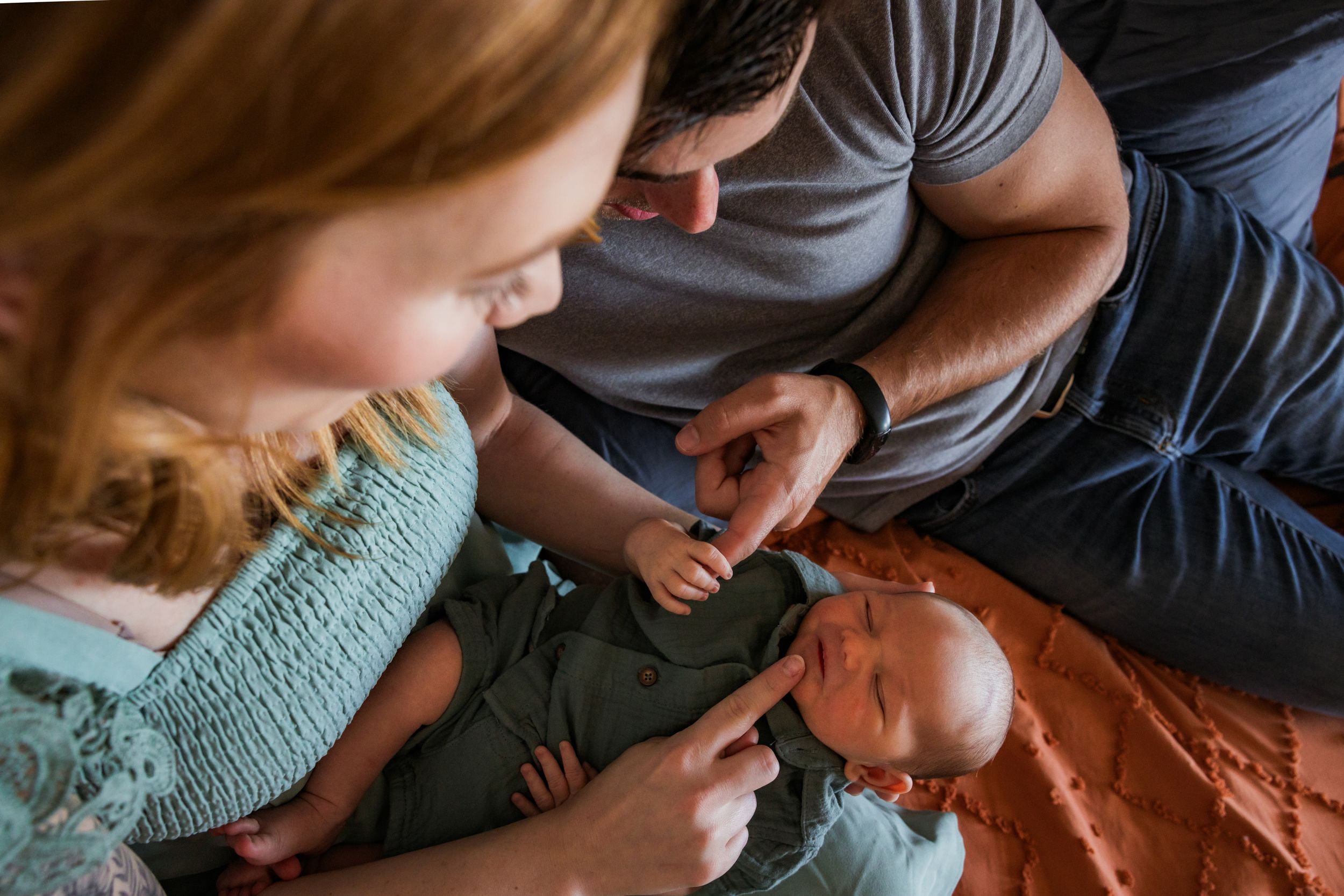 A couple sits close together on a bed while looking tenderly at newborn baby.