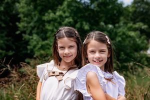 Two young girls in white blouses smile back to back while standing outdoors in a natural setting.