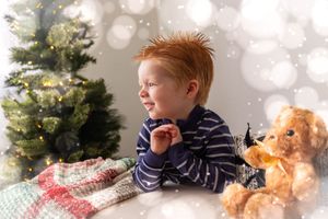 Red-haired toddler boy smiling near Christmas tree with stuffed bear toy and snowy bokeh background.