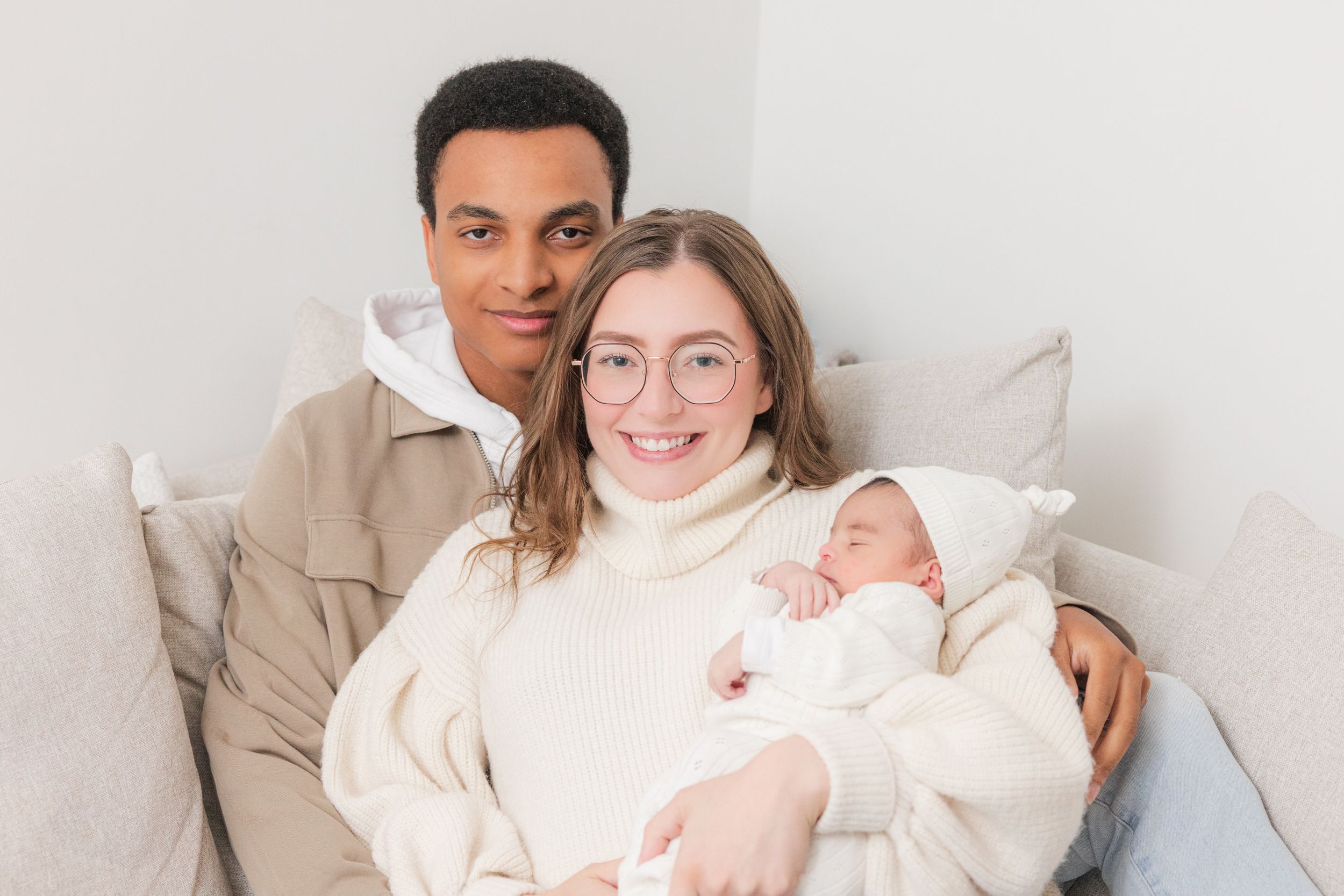 Happy couple sitting on a couch smiling while holding their newborn baby wrapped in a white blanket.