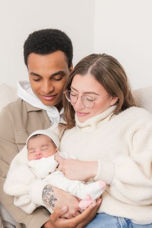 A couple in cozy sweaters lovingly holds their sleeping newborn baby dressed in white.