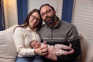 A smiling couple sits on a couch holding their newborn baby dressed in pink, posing for a family portrait.