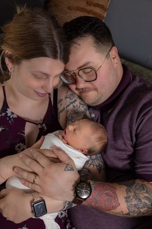 Two tattooed parents lovingly cradle their sleeping newborn baby, dressed in white, in a cozy home setting.