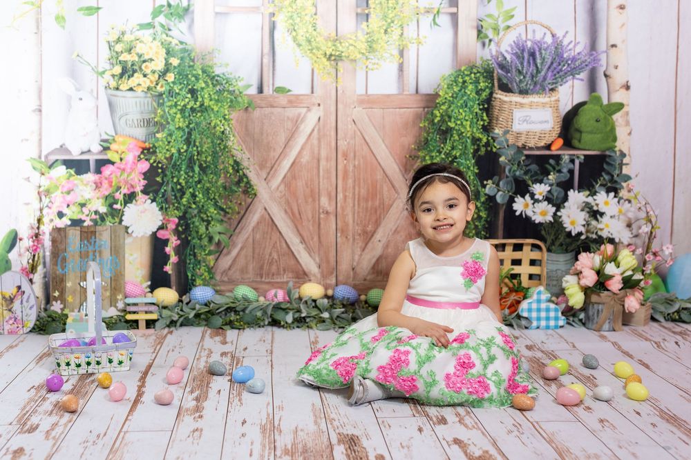 A child sits in a floral dress on whitewashed wooden floors against a backdrop of rustic barn doors and spring flowers.