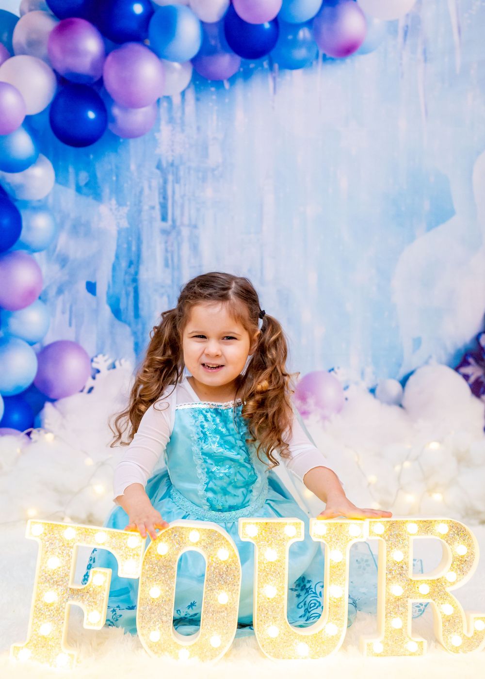 Cheerful child in a turquoise dress poses against a dreamy backdrop of blue and pink balloon decorations.