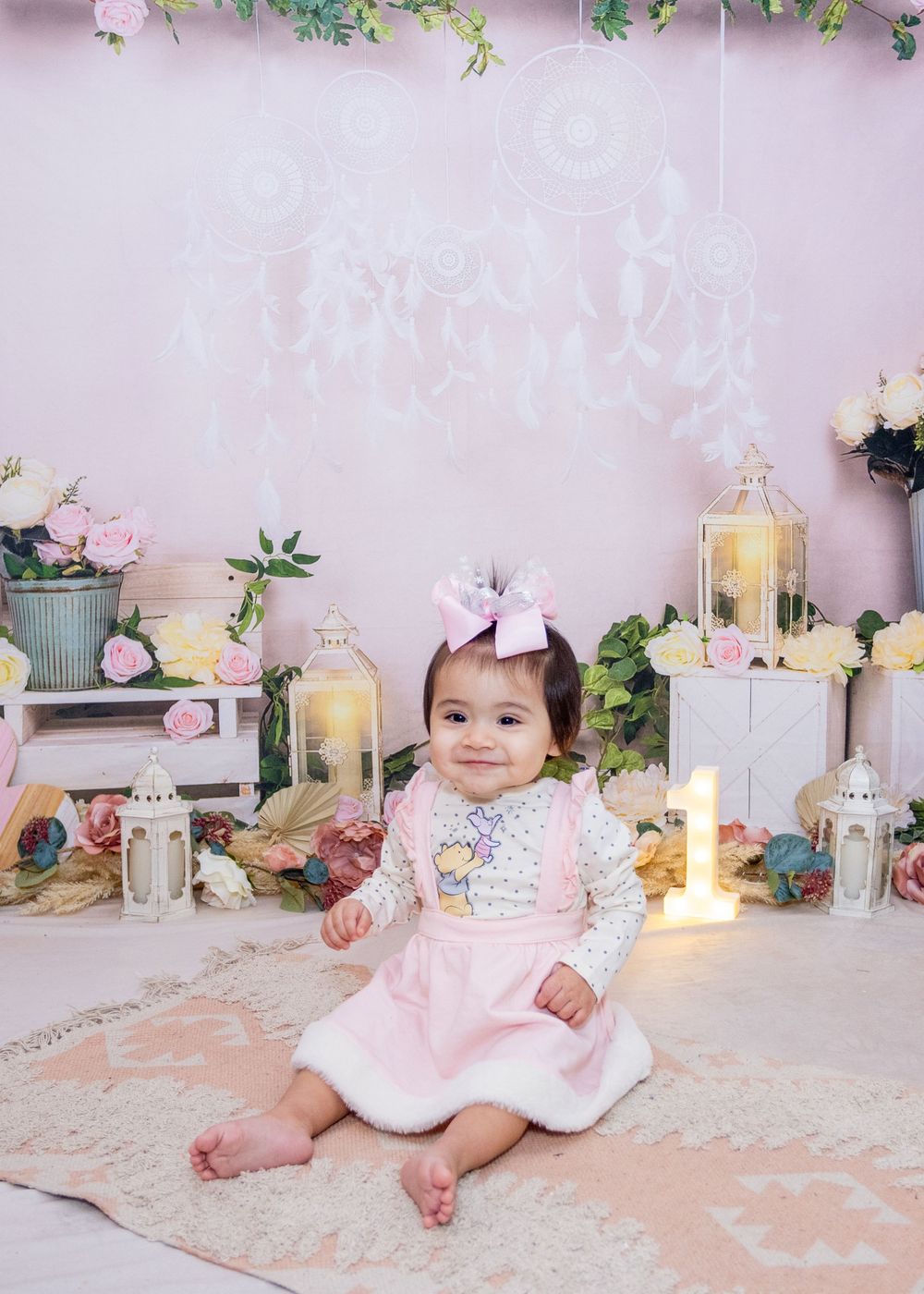 Baby in pink dress sitting on rug with decorative lanterns and flowers for photoshoot against pastel pink backdrop.