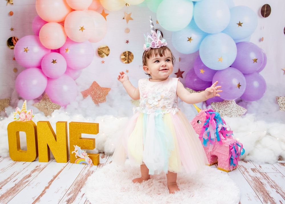 A festive scene with rainbow balloons, gold ONE letters, and a unicorn toy against a white backdrop.