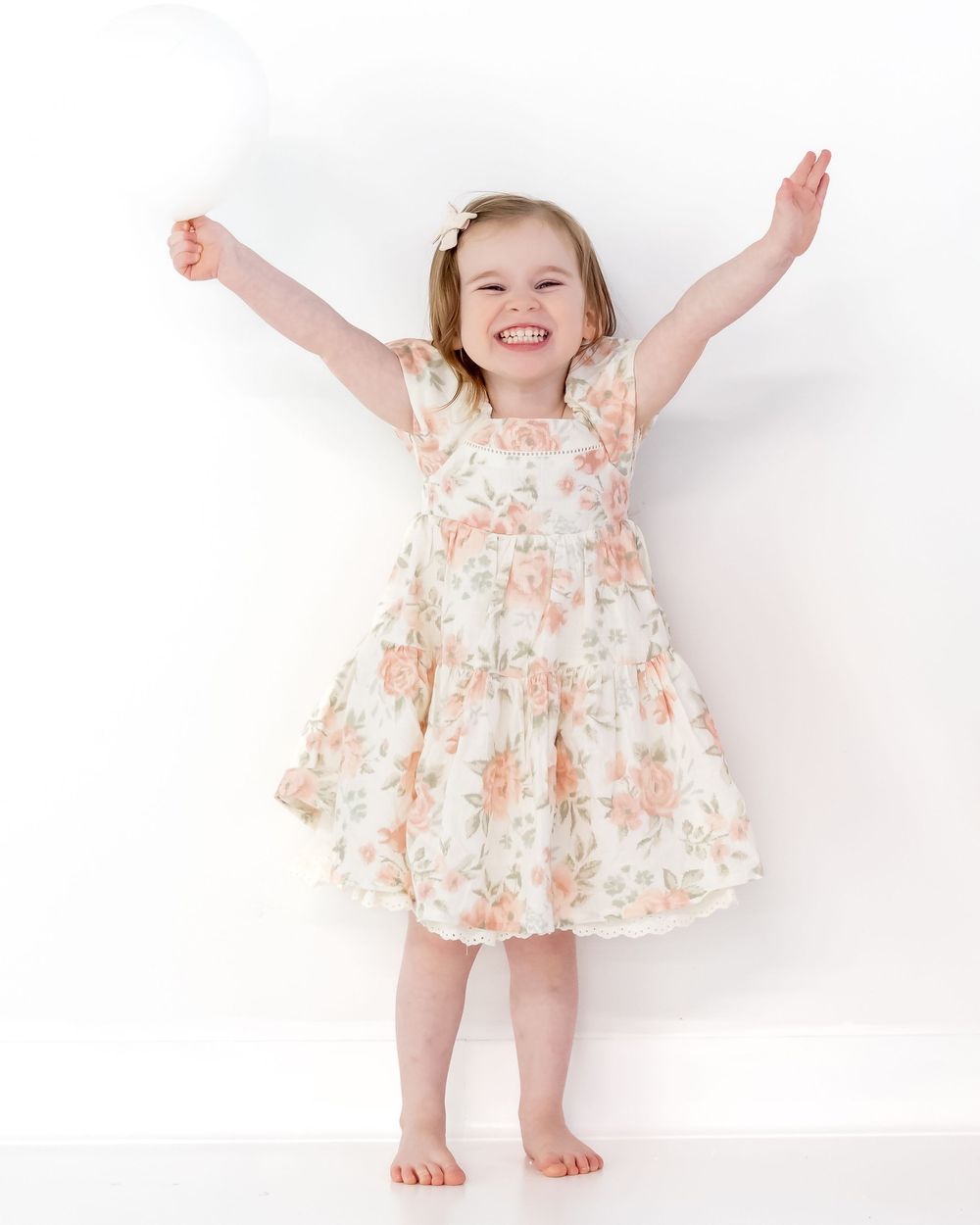 A child wearing a floral dress stands barefoot against a white background with arms raised joyfully.