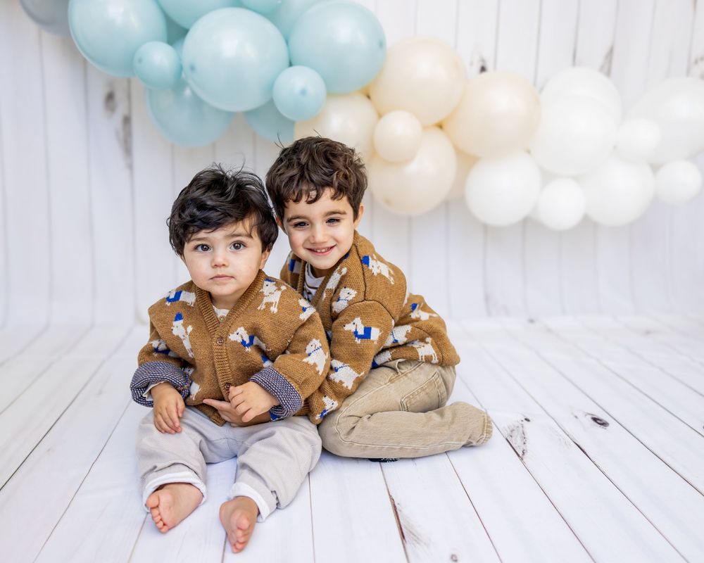 Two children wearing matching brown floral print jackets sit on white floor under blue and white balloon garland.