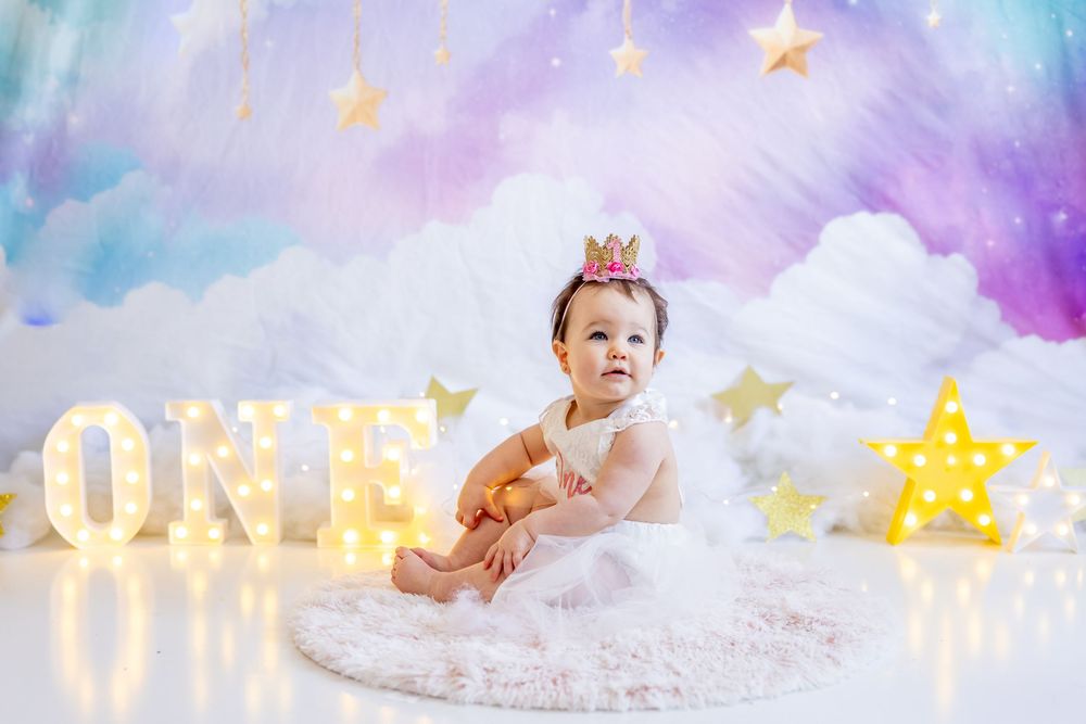 First birthday photo setup with illuminated ONE letters, clouds, stars, and soft pastel pink and purple background.