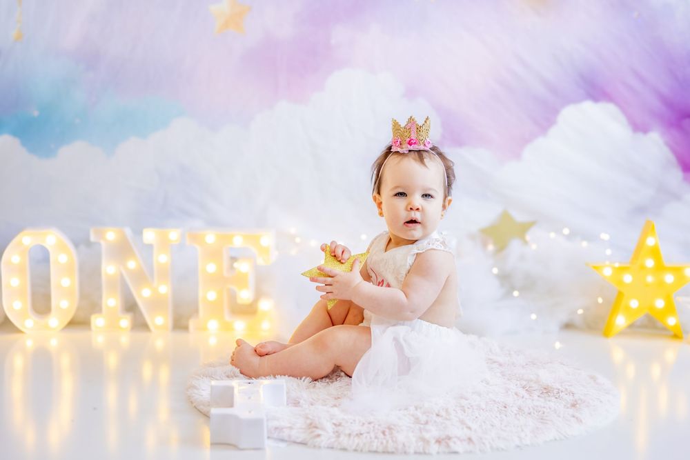 A baby in white dress sits near illuminated 'ONE' letters with pastel backdrop and decorative stars.