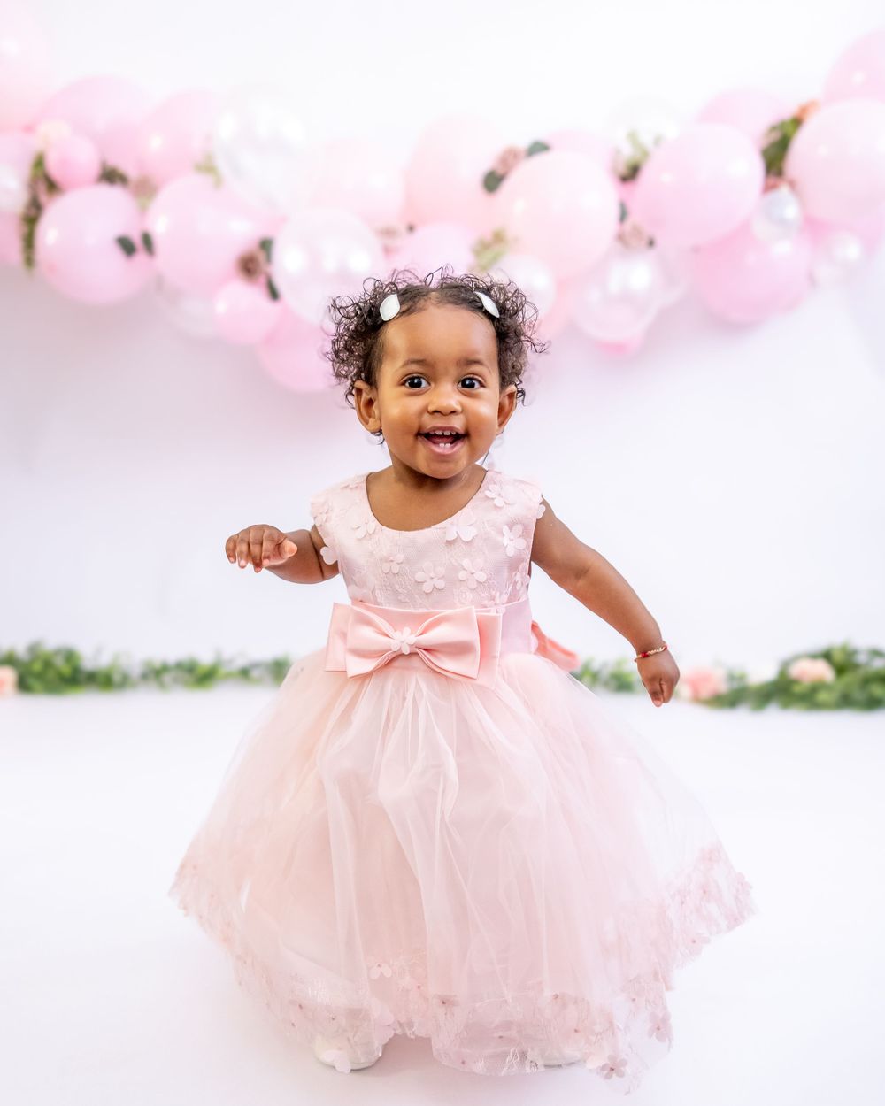 A toddler wearing a pink tulle party dress with a bow stands joyfully against a backdrop of pink balloon decorations.