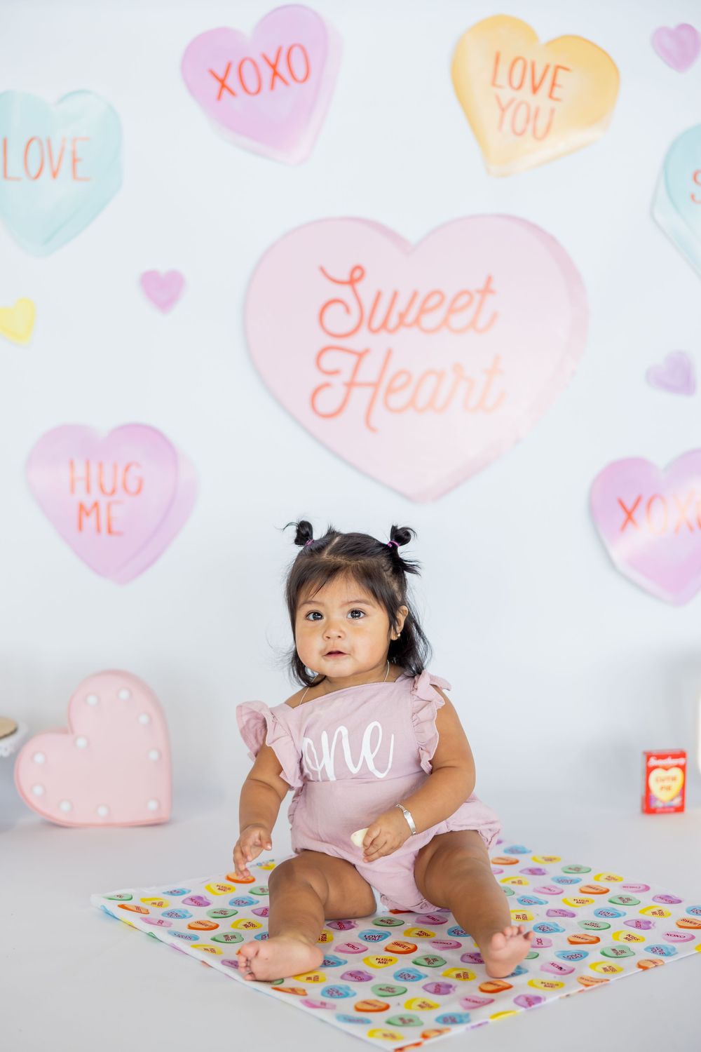 A small toddler in a pink outfit sits on a patterned mat against a wall decorated with pastel candy heart designs.