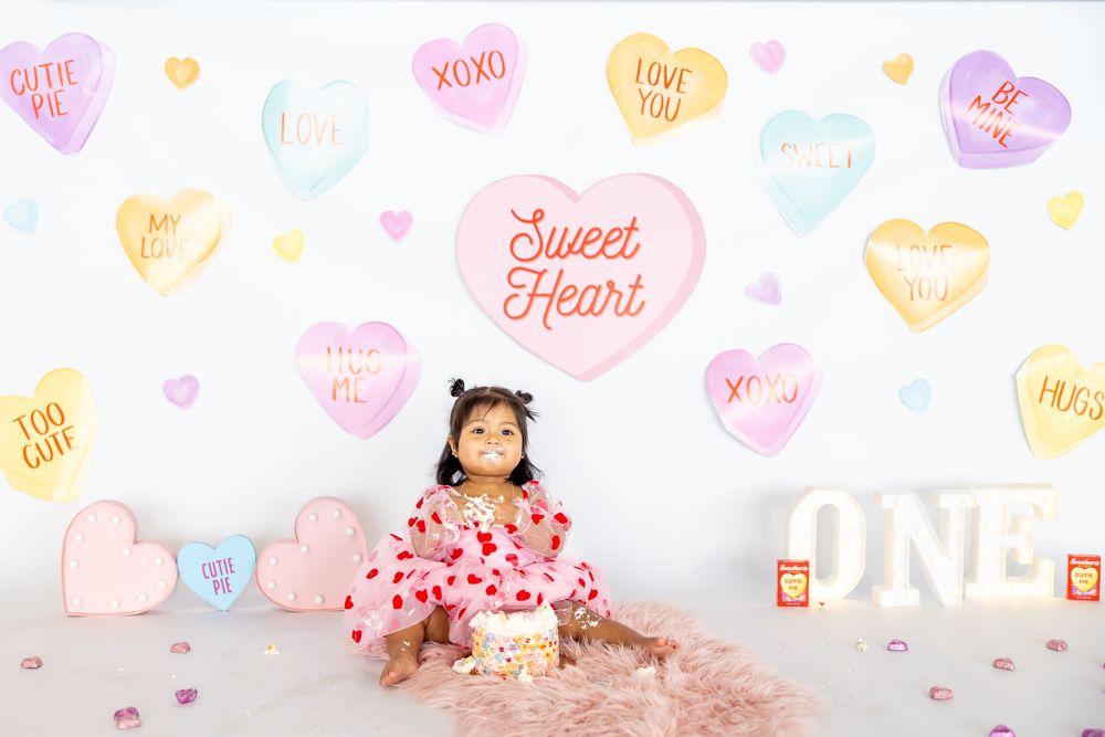 Baby's first birthday party setup with candy heart decorations, pink backdrop, and balloon arrangements against white wall.
