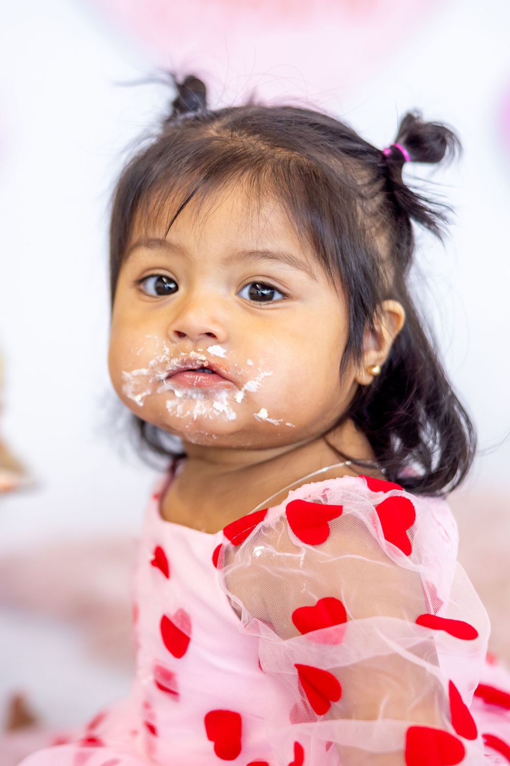 Little girl wearing pink dress with red hearts has messy frosting around mouth after eating dessert.