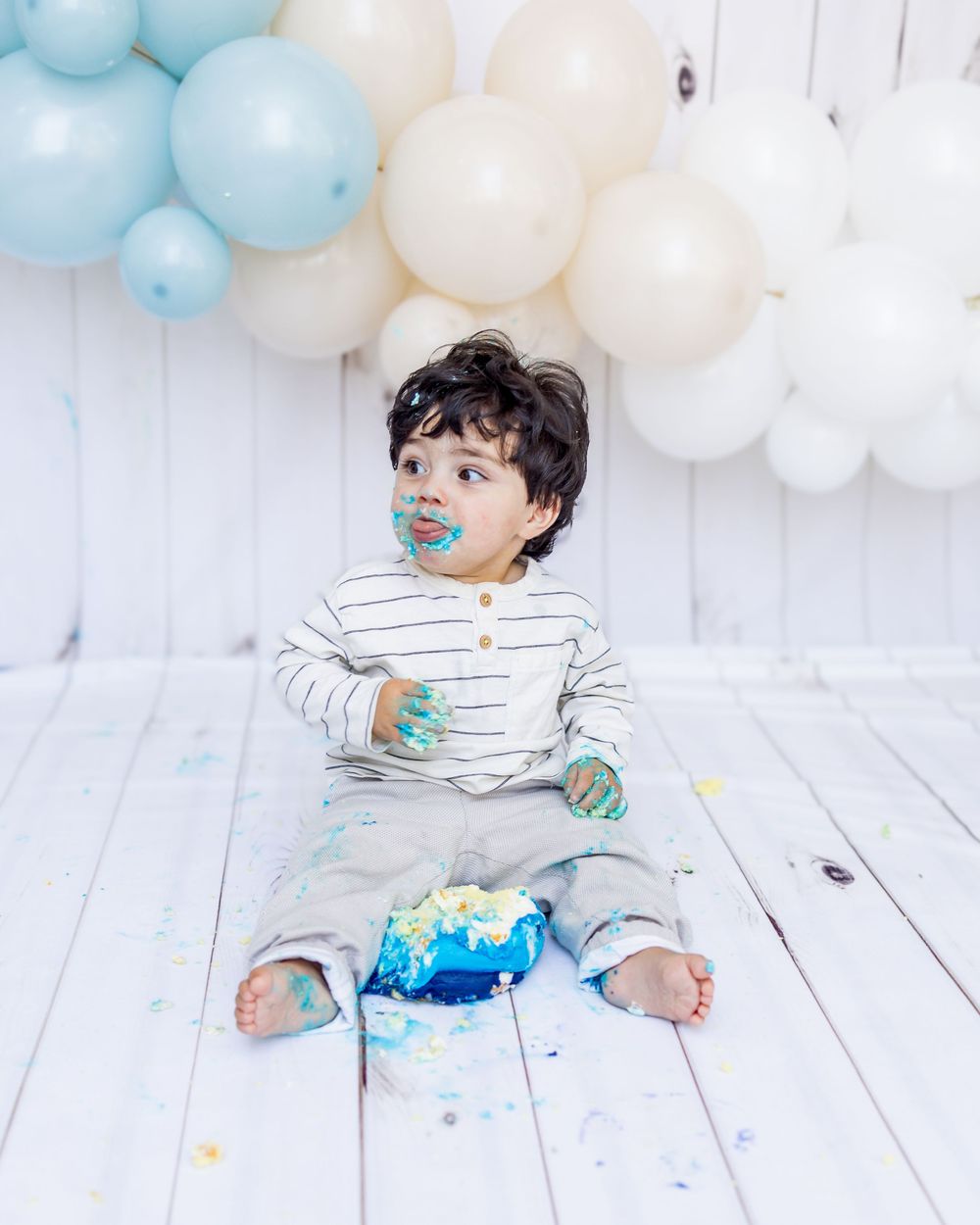 Young child in striped shirt sitting on white floor with blue and white balloon decorations and cake smash setup.