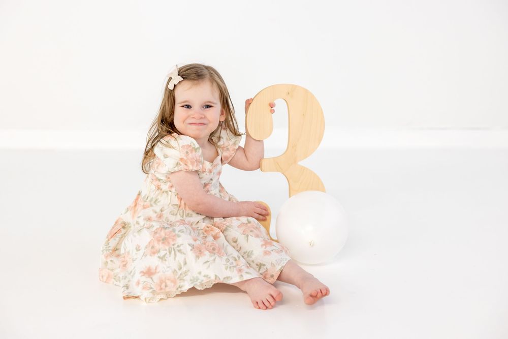 A young child in a floral dress sits beside a wooden number 3 and white balloon on a white background.