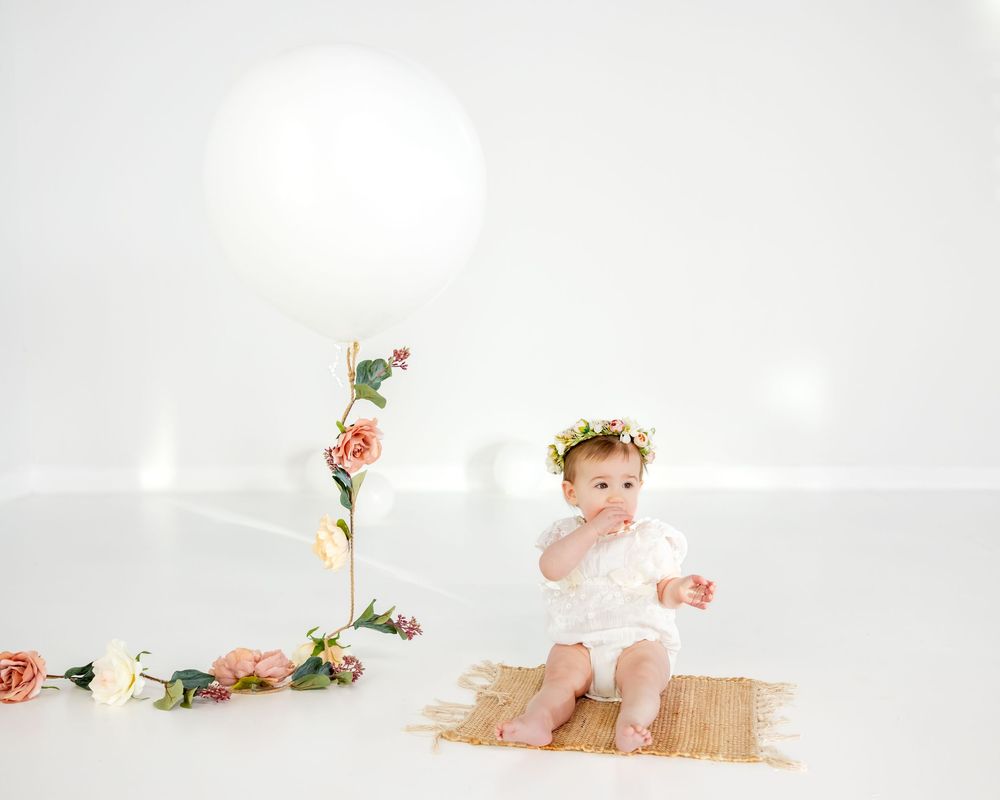 A floral garland decorates a white balloon next to a child in white dress and floral crown.