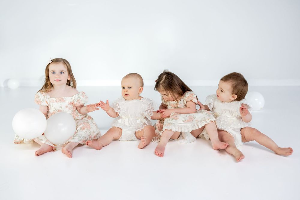 Four young children in white outfits sit and play together on a light gray backdrop.