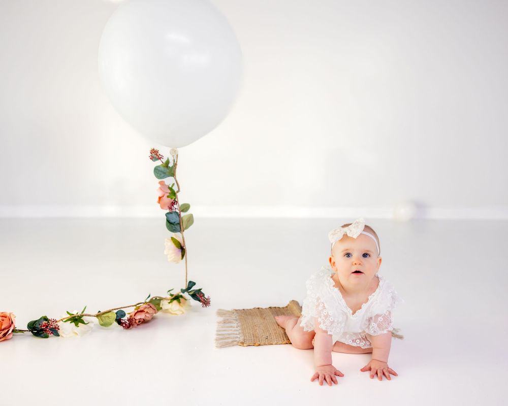 Baby in white outfit sits on floor next to large white balloon decorated with floral garland.