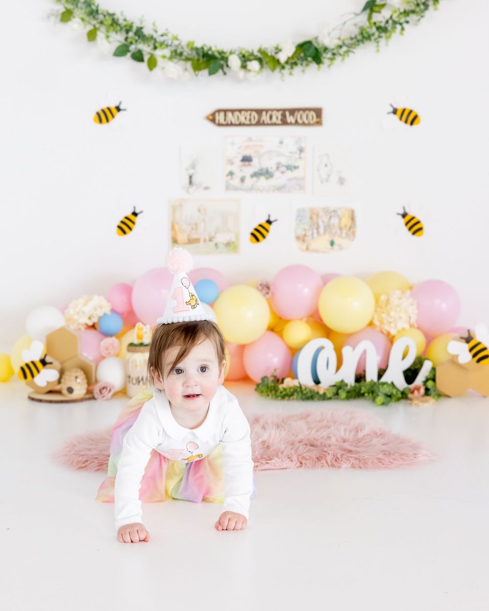 Sweet first birthday setup with pink and yellow balloon arch, bees, and leafy garland decorations against white backdrop.