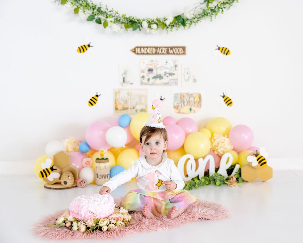 Pastel balloon display with honeybee decorations and floral garland creates whimsical first birthday setup on pink backdrop.