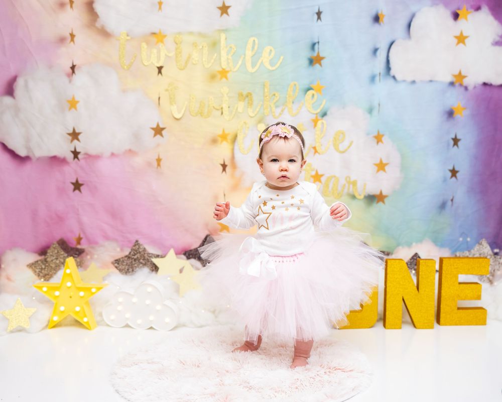 A baby in a white tutu dress stands against a dreamy pastel backdrop with gold stars and fluffy clouds.