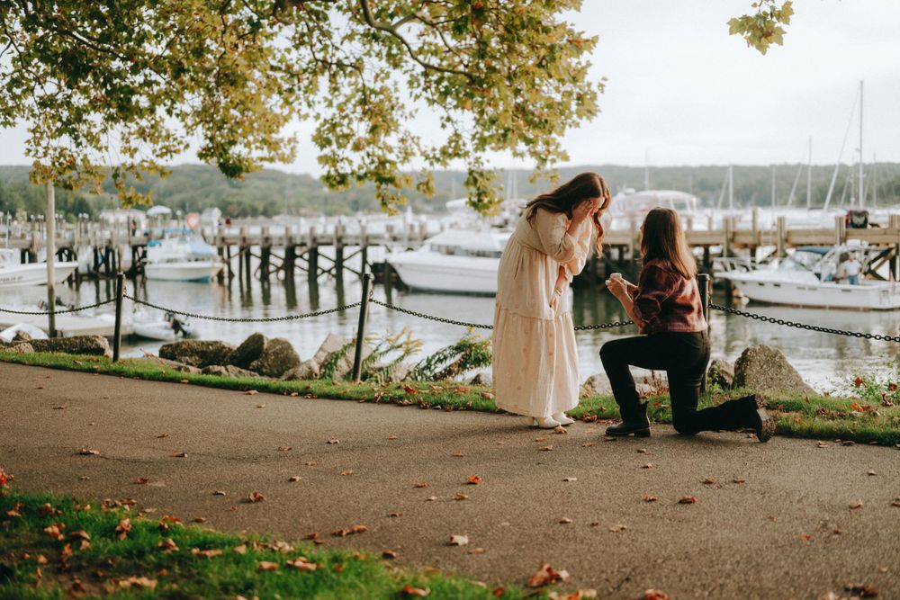 Romantic proposal scene by a marina with boats and autumn trees in background.
